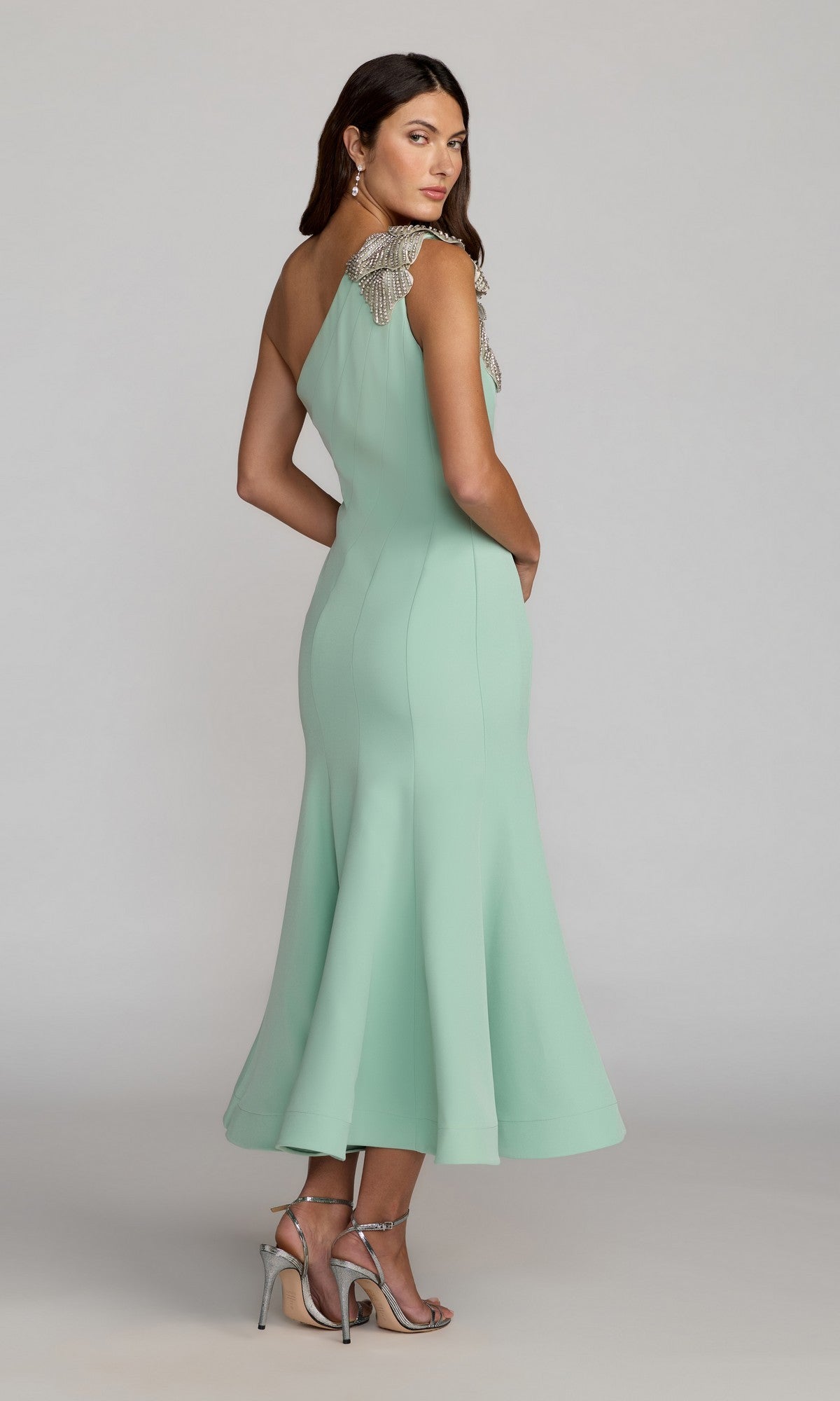 A woman stands in a neutral studio setting wearing the Tea-Length Party Dress: Mac Duggal 13221 in mint green crepe—an elegant one-shoulder midi dress accented with an embellished floral shoulder applique and silver strap details—paired with silver high-heeled sandals.